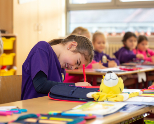 Ein Mädchen der Antoniusschule Oberachern sitzt lachend an ihrer Schulbank und schreibt mit einem roten Stift in ihr Heft.
